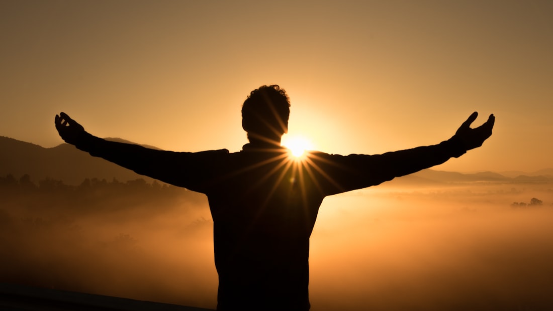 A person in silhouette with arms outstretched, facing a golden sunrise over misty hills
