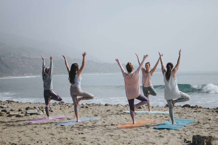 Five women in tree pose on colorful yoga mats along a misty beach, arms raised toward the ocean
