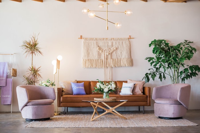 A warm, airy lounge with a tan leather sofa, mauve velvet chairs, potted plants, and a woven wall hanging