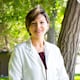 Smiling woman in a white coat standing outdoors in front of a tree with bright green leaves