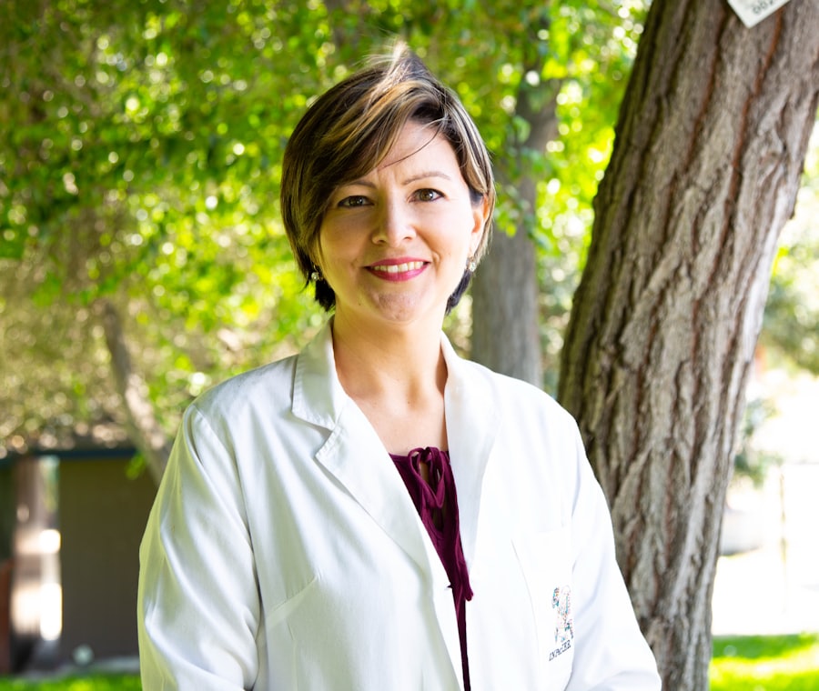 Smiling woman in a white coat standing outdoors in front of a tree with bright green leaves