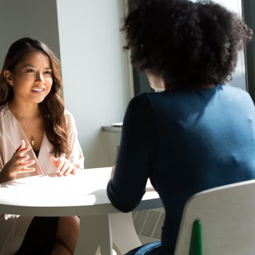 Two women seated across a small round table by a window, talking in a bright room