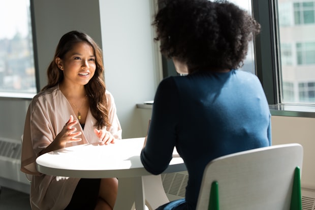 Two women seated across a small round table by a window, talking in a bright room