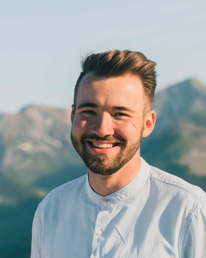 A smiling young bearded man in a light shirt outdoors, softly blurred mountains rising behind him